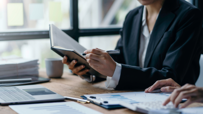 Professional in dark suit writing in notebook at desk with laptop and business documents.